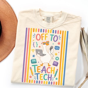 May include: A cream-colored t-shirt featuring a graphic of a white goose wearing rainbow heart-shaped sunglasses, holding a laptop and computer mouse. The text reads "OFF TO TEACH TECH" surrounded by colorful icons like pencils, keys, and stars.