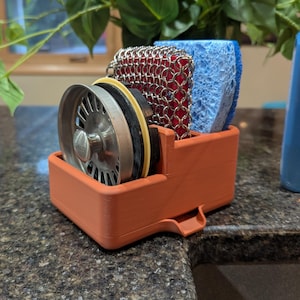 Peut inclure: Un organiseur d'évier orange imprimé en 3D contient une passoire d'évier en acier inoxydable, une éponge bleue et un récureur en cotte de mailles rouge. Une bouteille bleue est en arrière-plan. Le support est sur un comptoir sombre.