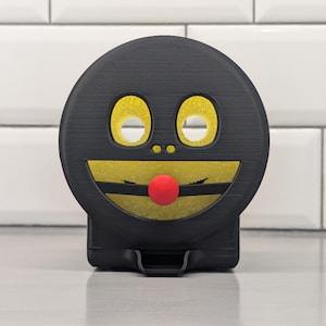May include: A black, round tape dispenser with a smiling face design. It has yellow eyes, a yellow and black smile, and a red nose. The dispenser has a textured surface and a small base. It is designed to hold tape.