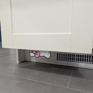 Op de afbeelding: Een witte kast met een roze en witte striksticker aan de onderkant. De strik is een cartoonontwerp. De kast staat boven een ventilatieopening met een grijze vloer op de voorgrond.
