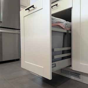 Puede incluir: Armario de cocina blanco con cubo de basura extraíble. La puerta del armario está abierta, revelando un cubo de basura gris y un estante. Tiradores negros y una rejilla de ventilación visibles. El suelo es de baldosas.