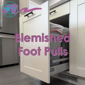 Op de afbeelding: Witte keukenkast met een uittrekbare lade. De lade is gedeeltelijk open en onthult een grijze vuilnisbak. De kast heeft zwarte handgrepen en de tekst "Blemished Foot Pulls" in roze.