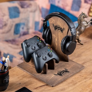May include: A wooden headphone stand with a pair of black gaming controllers and a set of black headphones. The stand has the text "Gamer's" and a controller graphic. A pen holder and a bronze horse statue are also visible.