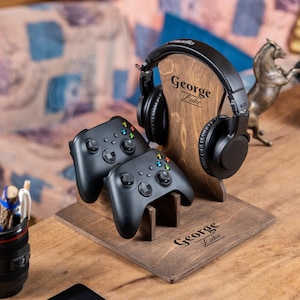 Puede incluir: Un soporte de madera para auriculares y mandos de juego con el texto grabado "George Luke". Dos mandos de juego negros y unos auriculares negros se muestran en el soporte. También se ve un portalápices y una pequeña estatua de caballo.