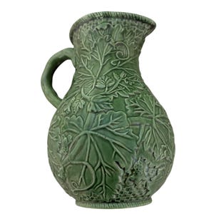 May include: Green ceramic pitcher with a handle and spout. The pitcher is decorated with a raised relief pattern of grape leaves and vines. The pitcher has a rounded body and a textured rim.