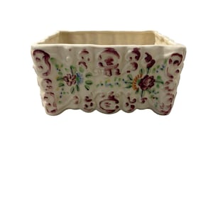 以下が含まれることがあります： クリーム色の長方形プランター、縁はスカラップ。プランターは、バーガンディ、青、黄色の花柄とスクロールワークのデザインで装飾されています。内側は明るいベージュ色です。