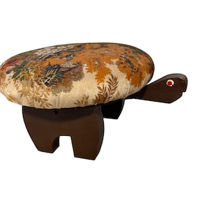 May include: A turtle-shaped footstool with a dark brown wooden base and a round, cushioned top upholstered in a cream-colored fabric featuring a detailed, multi-colored floral and foliage pattern. The head has a small, circular red eye detail.