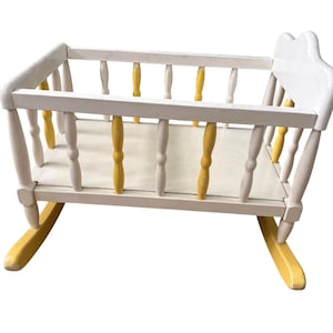 May include: A wooden doll cradle featuring a white frame with alternating yellow and white vertical spindles. The cradle has a flat white base and two curved yellow rockers at the bottom, with a solid white headboard at one end of the structure.