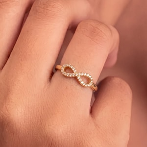 Puede incluir: Un anillo de tono dorado con un símbolo de infinito como pieza central. El bucle del infinito está incrustado con pequeñas piedras claras y brillantes en toda su superficie, creando un efecto continuo y resplandeciente en el anillo.