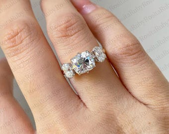 Puede incluir: Un anillo de oro con tres piedras preciosas claras. La piedra central es de talla cuadrada, flanqueada por dos piedras de talla ovalada. El anillo se muestra en una mano, resaltando el brillo. Es una joya.