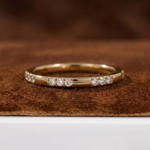 Op de afbeelding: Een gouden bandring met een reeks kleine, heldere edelstenen rond de band. De ring heeft een gepolijste afwerking en wordt gepresenteerd op een bruin oppervlak. De ring is een delicaat sieraad.