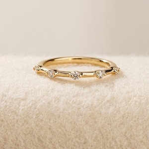 May include: A thin gold band ring featuring a series of small, round, clear stones set at regular intervals along the outer circumference. The metal has a polished, reflective finish, and the stones are secured in place by small metal prongs.
