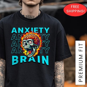 May include: Black t-shirt with a graphic design featuring a skull with a brain and the words "ANXIETY BRAIN" in blue and white. The design has a Halloween theme. The shirt is a premium fit.