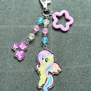 May include: A clear acrylic keychain featuring a yellow cartoon pony with a rainbow mane and tail, and pink wings. The keychain has a silver clasp, a pink star charm, and a beaded strand with pink, blue, and yellow beads, and pink butterfly charms.