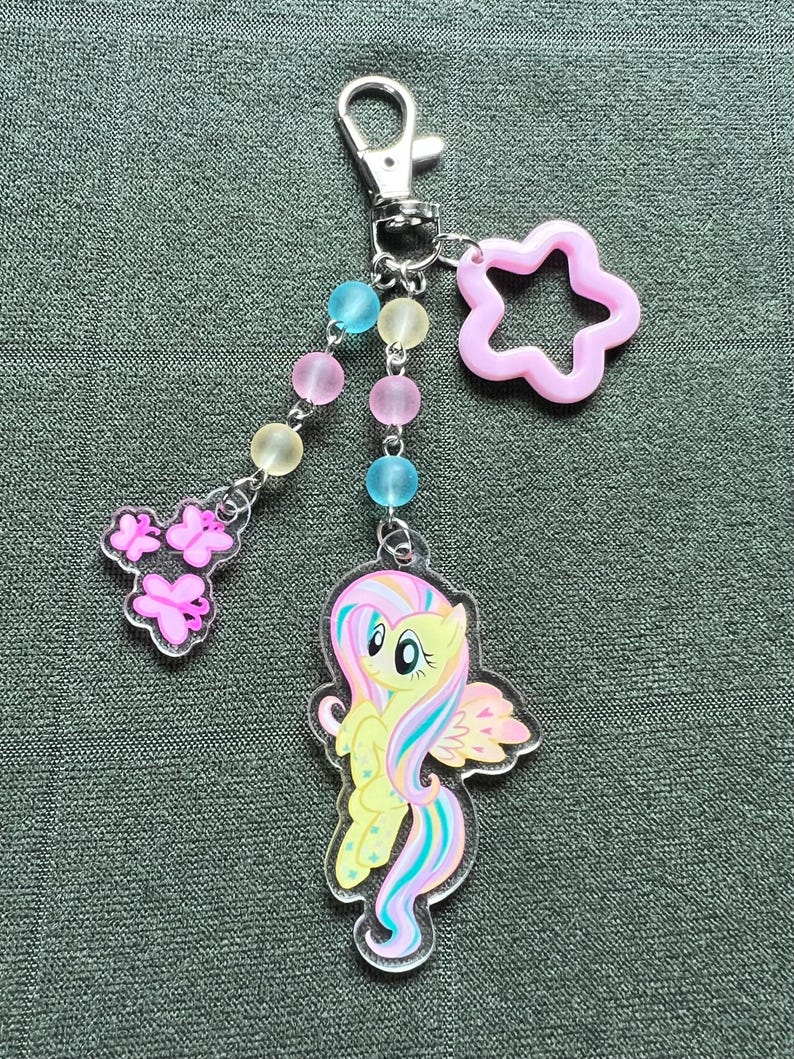May include: A clear acrylic keychain featuring a yellow cartoon pony with a rainbow mane and tail, and pink wings. The keychain has a silver clasp, a pink star charm, and a beaded strand with pink, blue, and yellow beads, and pink butterfly charms.