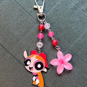 May include: A keychain featuring a cartoon character with a red bow and orange hair, a pink flower charm, and a beaded strand with red, pink, and clear beads. The keychain has a silver clasp.
