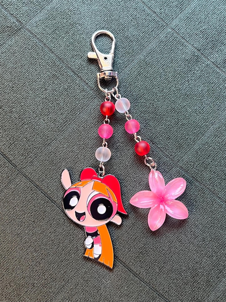 May include: A keychain featuring a cartoon character with a red bow and orange hair, a pink flower charm, and a beaded strand with red, pink, and clear beads. The keychain has a silver clasp.