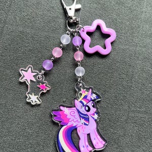 May include: A My Little Pony keychain featuring a purple pony with a rainbow tail and wings. The keychain includes a star-shaped charm, a star-shaped charm with stars, and a beaded chain with pink, purple, and white beads. The keychain is attached to a silver clasp.