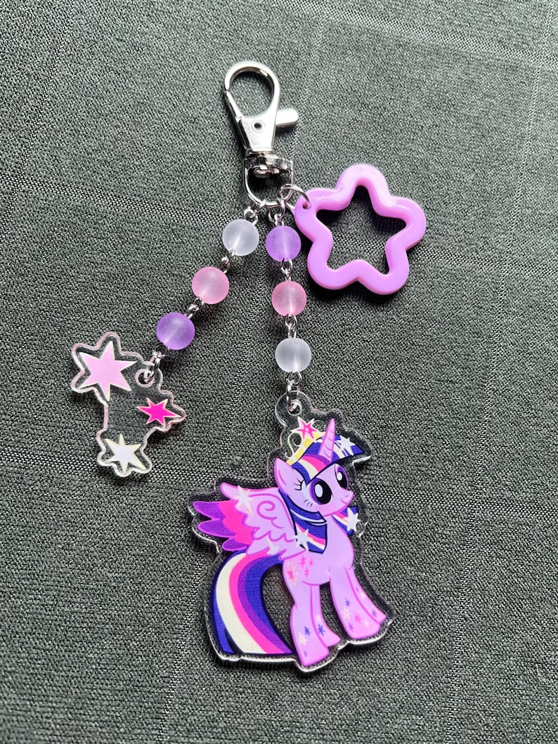 May include: A My Little Pony keychain featuring a purple pony with a rainbow tail and wings. The keychain includes a star-shaped charm, a star-shaped charm with stars, and a beaded chain with pink, purple, and white beads. The keychain is attached to a silver clasp.
