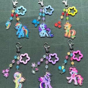 May include: A collection of six keychains featuring colorful cartoon pony designs. Each keychain has a silver clasp, a beaded strand, and a star-shaped charm. The pony charms are in various colors, including blue, pink, yellow, and purple.
