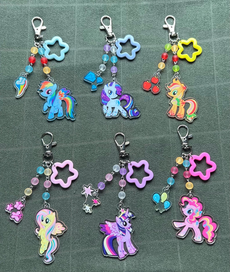May include: A collection of six keychains featuring colorful cartoon pony designs. Each keychain has a silver clasp, a beaded strand, and a star-shaped charm. The pony charms are in various colors, including blue, pink, yellow, and purple.