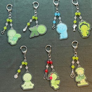 May include: A collection of keychains with various designs. Each keychain features a cartoon figure in different poses, some with laptops or musical notes. The keychains have silver-colored hardware and beaded accents in green, blue, and red.