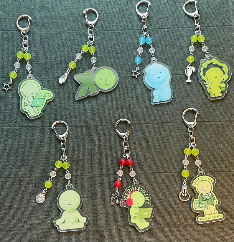 May include: A collection of keychains with various designs. Each keychain features a cartoon figure in different poses, some with laptops or musical notes. The keychains have silver-colored hardware and beaded accents in green, blue, and red.