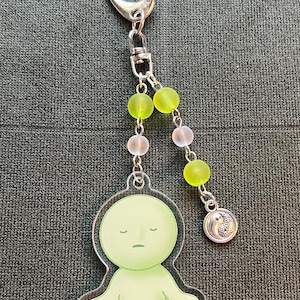 May include: A keychain featuring a light green figure in a meditative pose. The figure is attached to a silver chain with green and pink beads, and a silver charm with a yin-yang symbol. The keychain has a silver clasp.