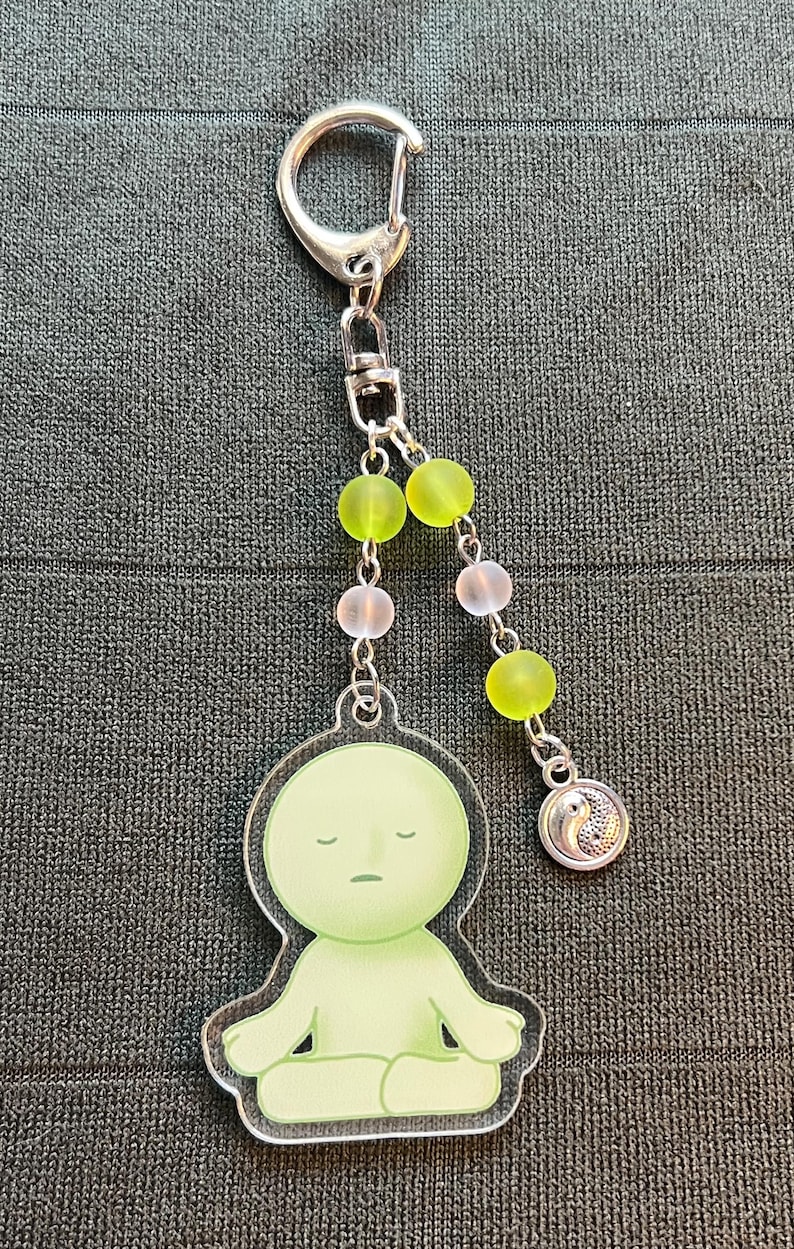 May include: A keychain featuring a light green figure in a meditative pose. The figure is attached to a silver chain with green and pink beads, and a silver charm with a yin-yang symbol. The keychain has a silver clasp.