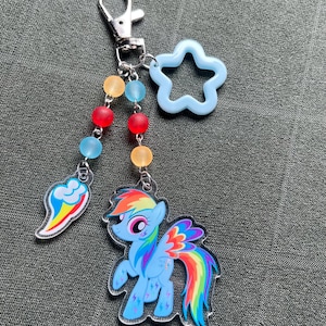 May include: A keychain featuring a cartoon pony with a rainbow mane and tail, a rainbow cloud, and a light blue star-shaped charm. The keychain has a silver clasp and colorful beads.