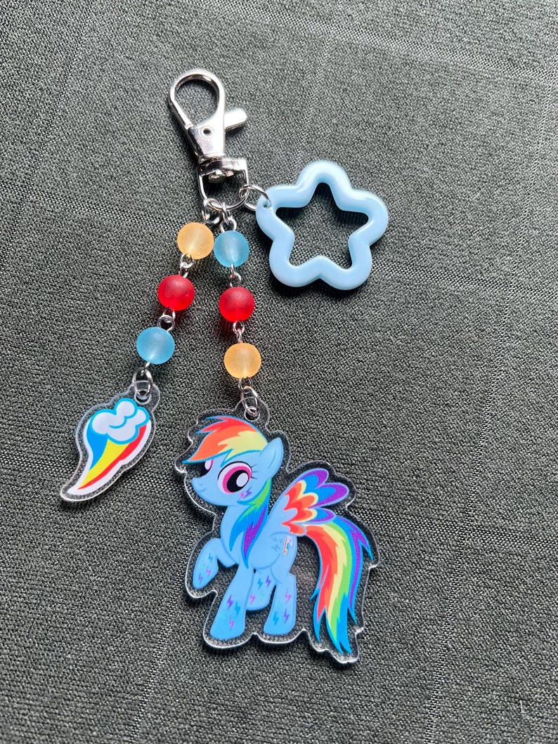 May include: A keychain featuring a cartoon pony with a rainbow mane and tail, a rainbow cloud, and a light blue star-shaped charm. The keychain has a silver clasp and colorful beads.