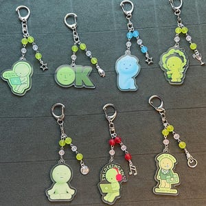 May include: Collection of keychains with various designs. Each keychain features a cartoon figure or text, such as "OK" or "I'm busy go away." The keychains have silver-colored rings and beaded accents in colors like green, red, and blue.