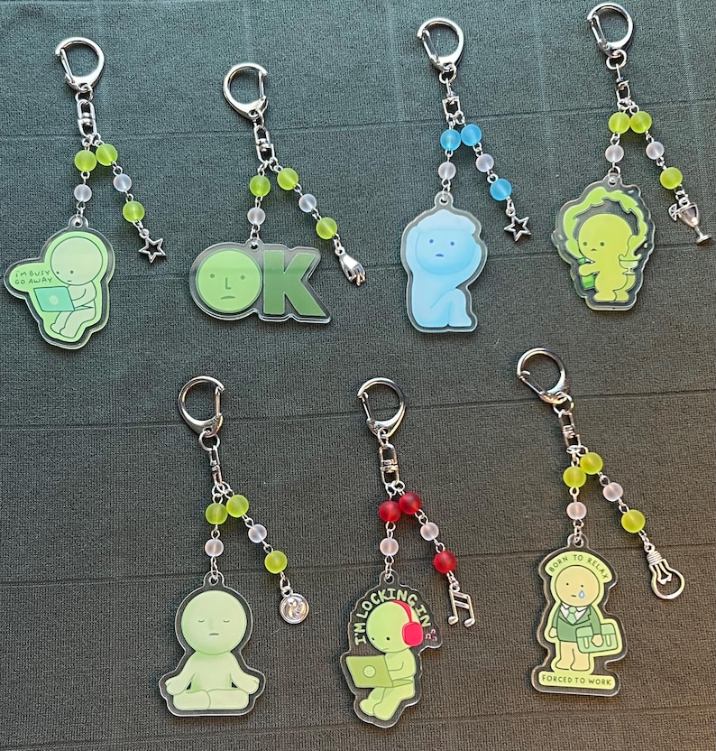 May include: Collection of keychains with various designs. Each keychain features a cartoon figure or text, such as "OK" or "I'm busy go away." The keychains have silver-colored rings and beaded accents in colors like green, red, and blue.