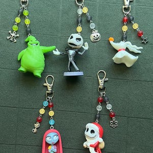 May include: Five character keychains featuring characters from The Nightmare Before Christmas. Each keychain has a silver clasp, a beaded strand with various colors, and a character charm. Characters include Oogie Boogie, Jack Skellington, Zero, Sally, and Jack dressed as Santa.