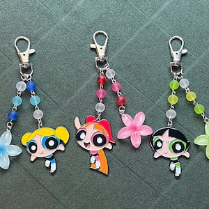 May include: Three keychains featuring cartoon characters with flower charms and beaded accents. Each keychain has a silver clasp and a different character design: a blonde, a redhead, and a black-haired character. The charms are in blue, pink, and green.