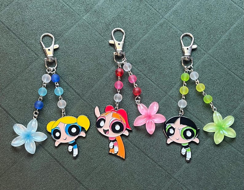 May include: Three keychains featuring cartoon characters with flower charms and beaded accents. Each keychain has a silver clasp and a different character design: a blonde, a redhead, and a black-haired character. The charms are in blue, pink, and green.