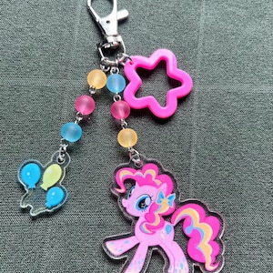 May include: A pink and clear acrylic keychain featuring a cartoon pony with a star-shaped charm and a charm with balloons. The keychain has a silver clasp and a beaded chain with pink, yellow, and blue beads.