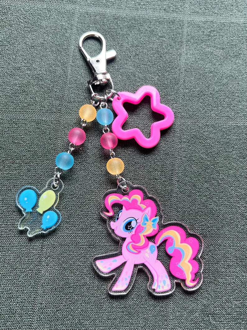 May include: A pink and clear acrylic keychain featuring a cartoon pony with a star-shaped charm and a charm with balloons. The keychain has a silver clasp and a beaded chain with pink, yellow, and blue beads.