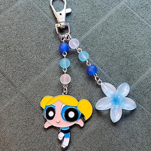 May include: A silver-tone keychain with a cartoon character charm, a flower charm, and a beaded strand. The cartoon character has yellow hair, blue eyes, and a blue and black outfit. The flower is light blue.