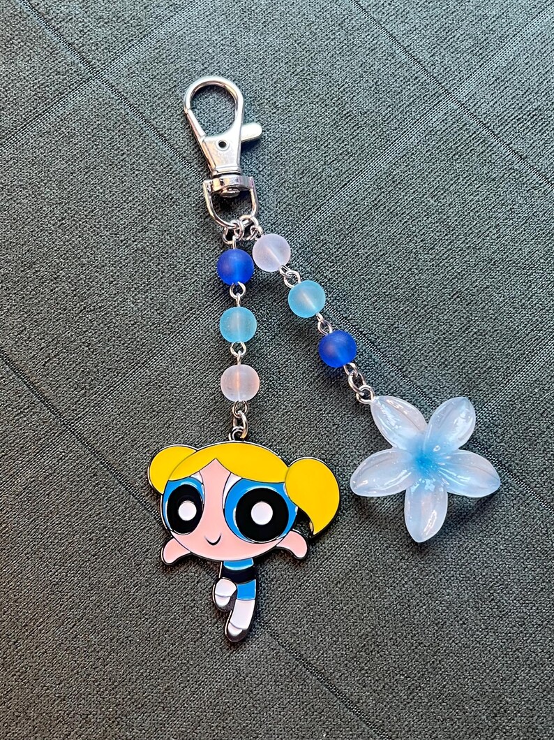 May include: A silver-tone keychain with a cartoon character charm, a flower charm, and a beaded strand. The cartoon character has yellow hair, blue eyes, and a blue and black outfit. The flower is light blue.