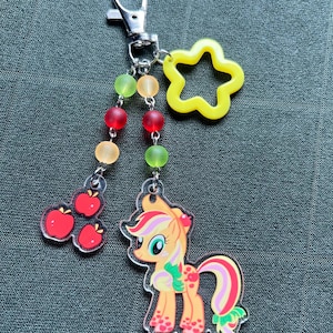 May include: A clear acrylic keychain featuring a cartoon pony with an apple theme, a yellow star, and red, orange, and green beads. The pony has an orange body, pink and green accents, and a red apple on its head. A cluster of red apples hangs from the chain.