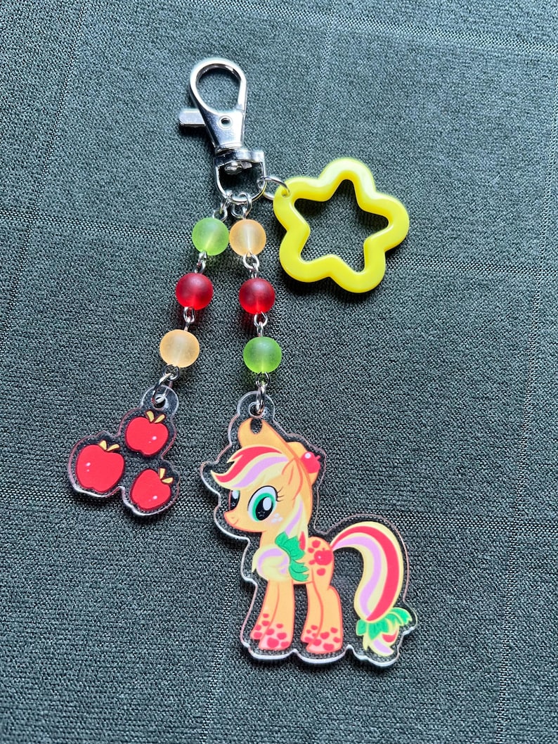 May include: A clear acrylic keychain featuring a cartoon pony with an apple theme, a yellow star, and red, orange, and green beads. The pony has an orange body, pink and green accents, and a red apple on its head. A cluster of red apples hangs from the chain.
