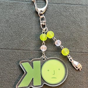 May include: A clear acrylic keychain with a green "OK" design and a cartoon face. The keychain has a silver clasp and is adorned with green, white, and pink beads, and a small silver hand charm.