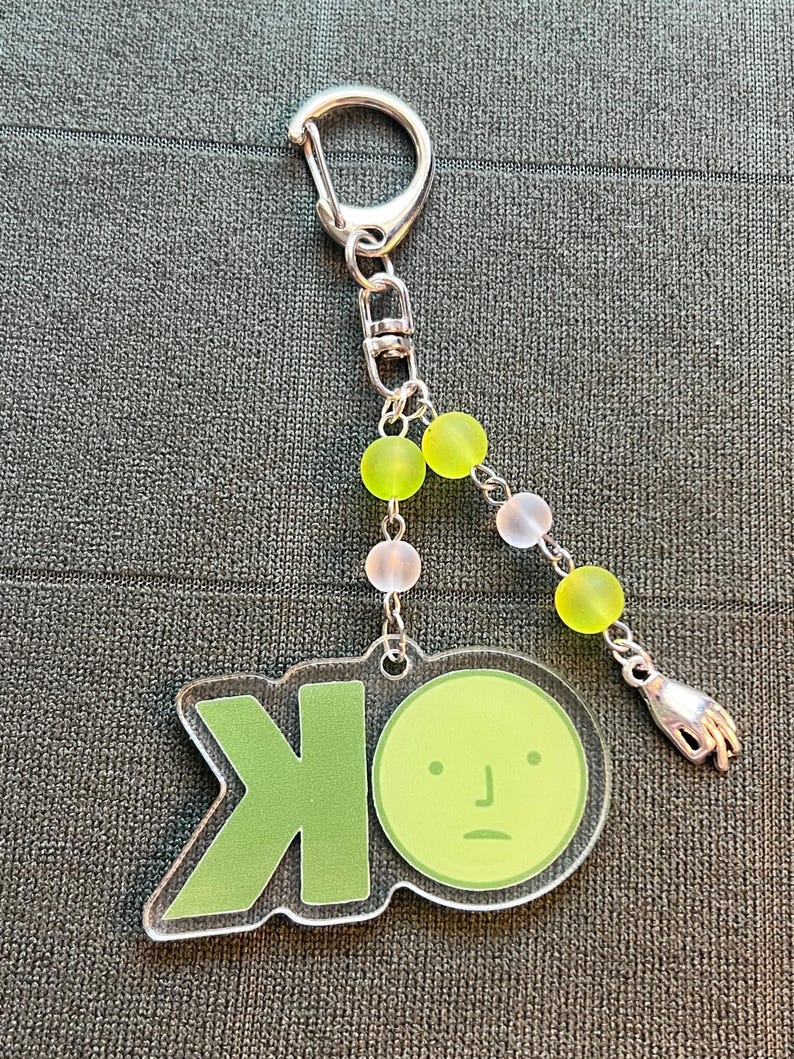 May include: A clear acrylic keychain with a green "OK" design and a cartoon face. The keychain has a silver clasp and is adorned with green, white, and pink beads, and a small silver hand charm.