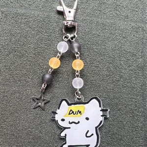 May include: A keychain with a silver clasp, featuring a white cat charm with the word "DUM" on a yellow sticky note. The keychain also includes gray, yellow, and clear beads, and a silver star charm.