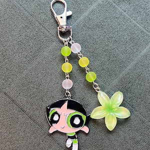 May include: A silver-tone keychain featuring a cartoon character, Buttercup, from The Powerpuff Girls. The charm is accented with green and white beads and a translucent green flower. The keychain has a lobster clasp.