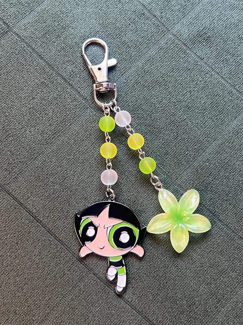 May include: A silver-tone keychain featuring a cartoon character, Buttercup, from The Powerpuff Girls. The charm is accented with green and white beads and a translucent green flower. The keychain has a lobster clasp.
