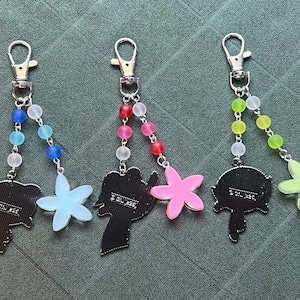 May include: Three keychains with silver clasps and beaded strands in blue, pink, and green. Each keychain features a charm: an elephant, a flower, and a stylized figure. The charms are in black, light blue, and pink.