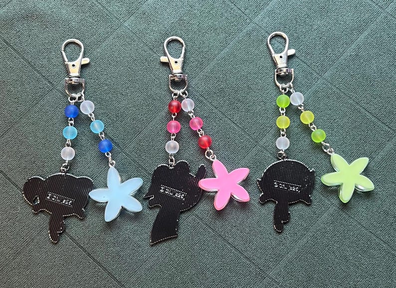 May include: Three keychains with silver clasps and beaded strands in blue, pink, and green. Each keychain features a charm: an elephant, a flower, and a stylized figure. The charms are in black, light blue, and pink.