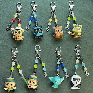 May include: A collection of character keychains with silver-tone clasps and beaded accents. The keychains feature various figures, including a bear-like creature, a green-skinned alien, and a character with a helmet. Each keychain has a unique design and color scheme.